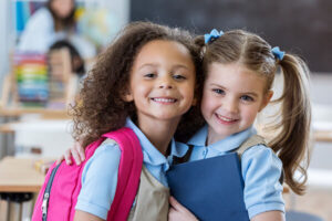 Adorable kindergarten or preschool students hug each other in their classroom. They are wearing school uniforms.